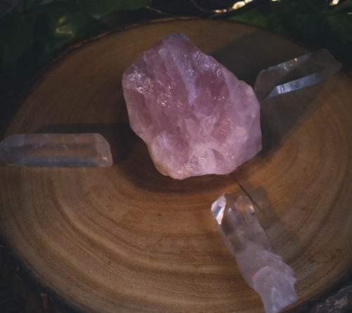 a simple crystal grid set up on a slice of wood. a large rose quartz sits in the center with 3 clear quartz radiating out from the center stone.